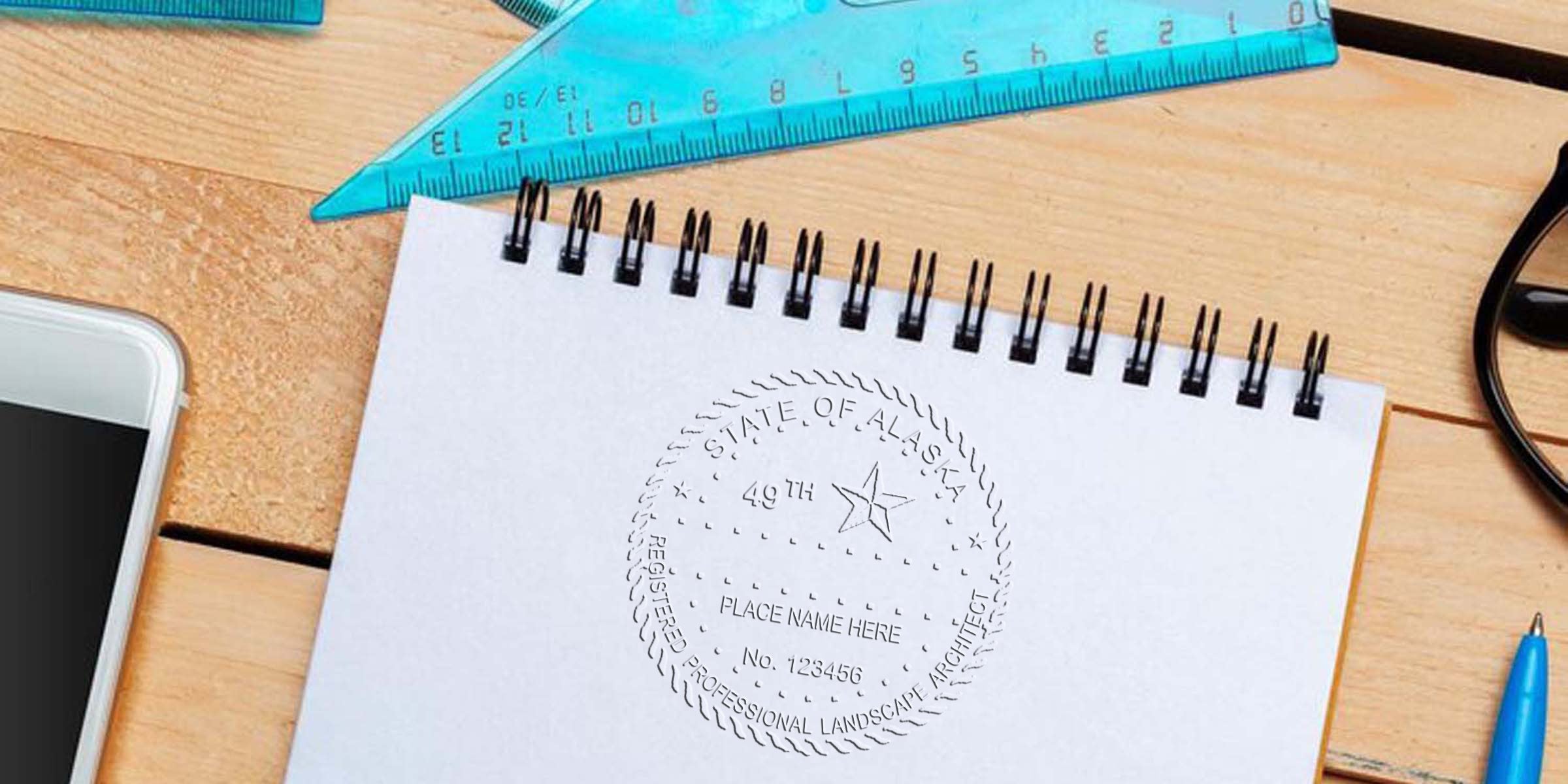 Alaska landscape architect stamp on white spiral notebook with blue ruler, smartphone, glasses, and pen on wooden desk, showing professional Alaska landscape architect license certification.