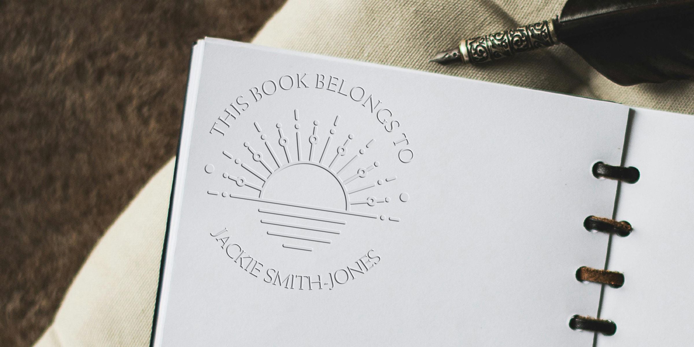 Embossed personalized book cover reading “This Book Belongs to Jackie Smith-Jones” with a decorative pen and bound notebook on a beige fabric background