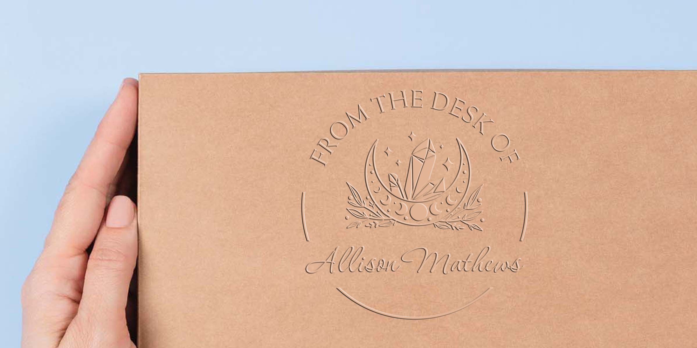Hand holding a personalized kraft gift box with embossed crystal moon logo and “From the Desk of Allison Mathews” branding on a light blue background