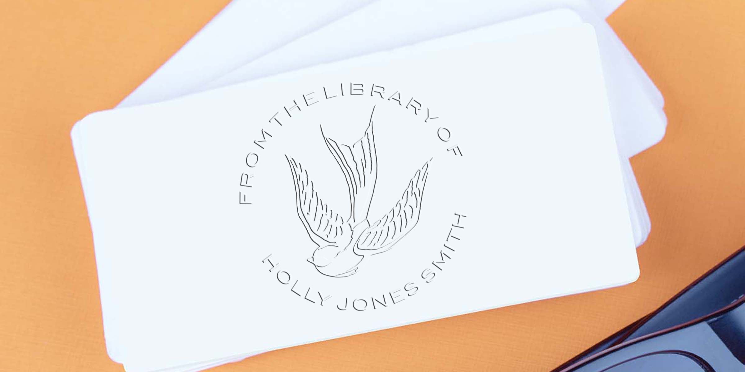 White bookplate cards with an embossed bird logo and text reading “From the Library of Holly Jones Smith” on an orange background with eyeglasses.
