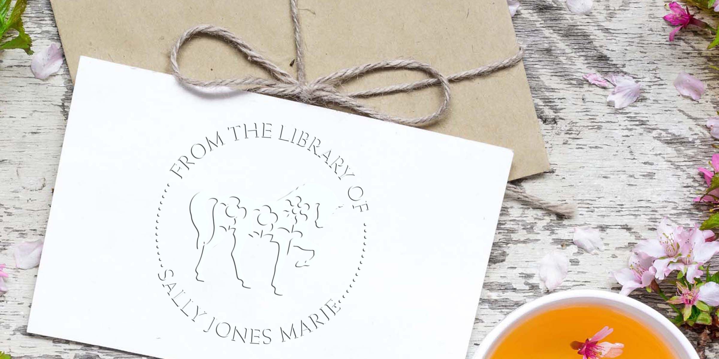 Custom library return stamp on white card tied to kraft paper with twine, surrounded by pink flower petals and tea on rustic wood background