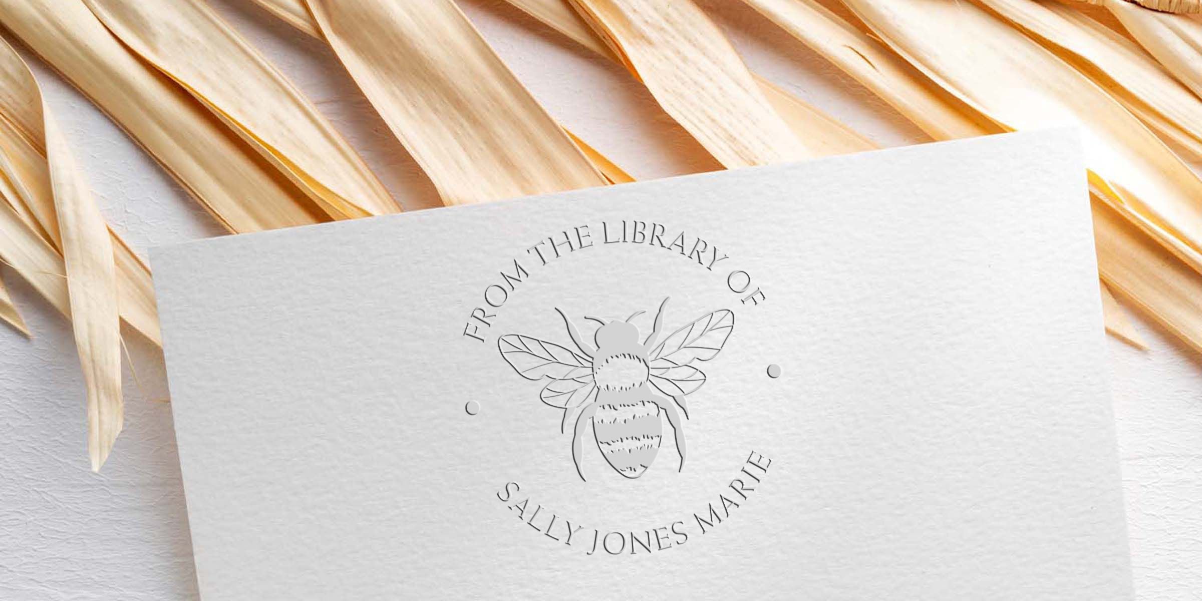 Embossed personalized library card with bee logo and “From the Library of Sally Jones Marie” text on a white card over dried palm leaves.
