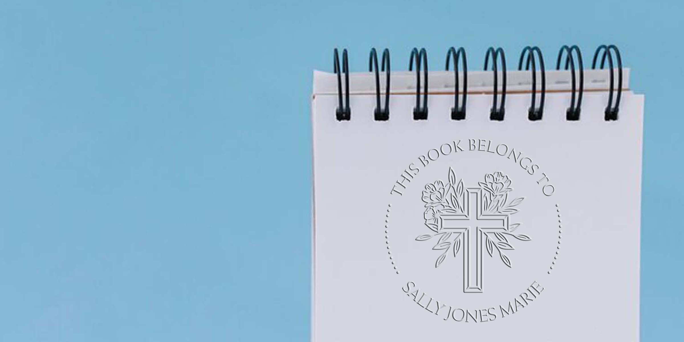 Personalized Christian spiral notebook with embossed cross and floral design, reading “This Book Belongs to Sally Jones Marie,” on a blue background