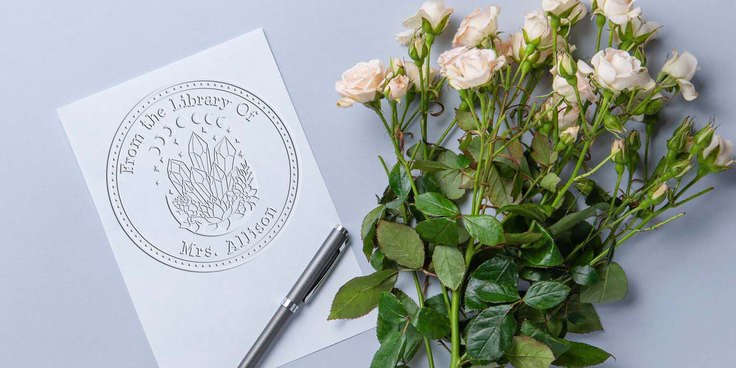 White roses beside a personalized “From the Library of Mrs. Allison” embossing seal design and silver pen on a light gray background