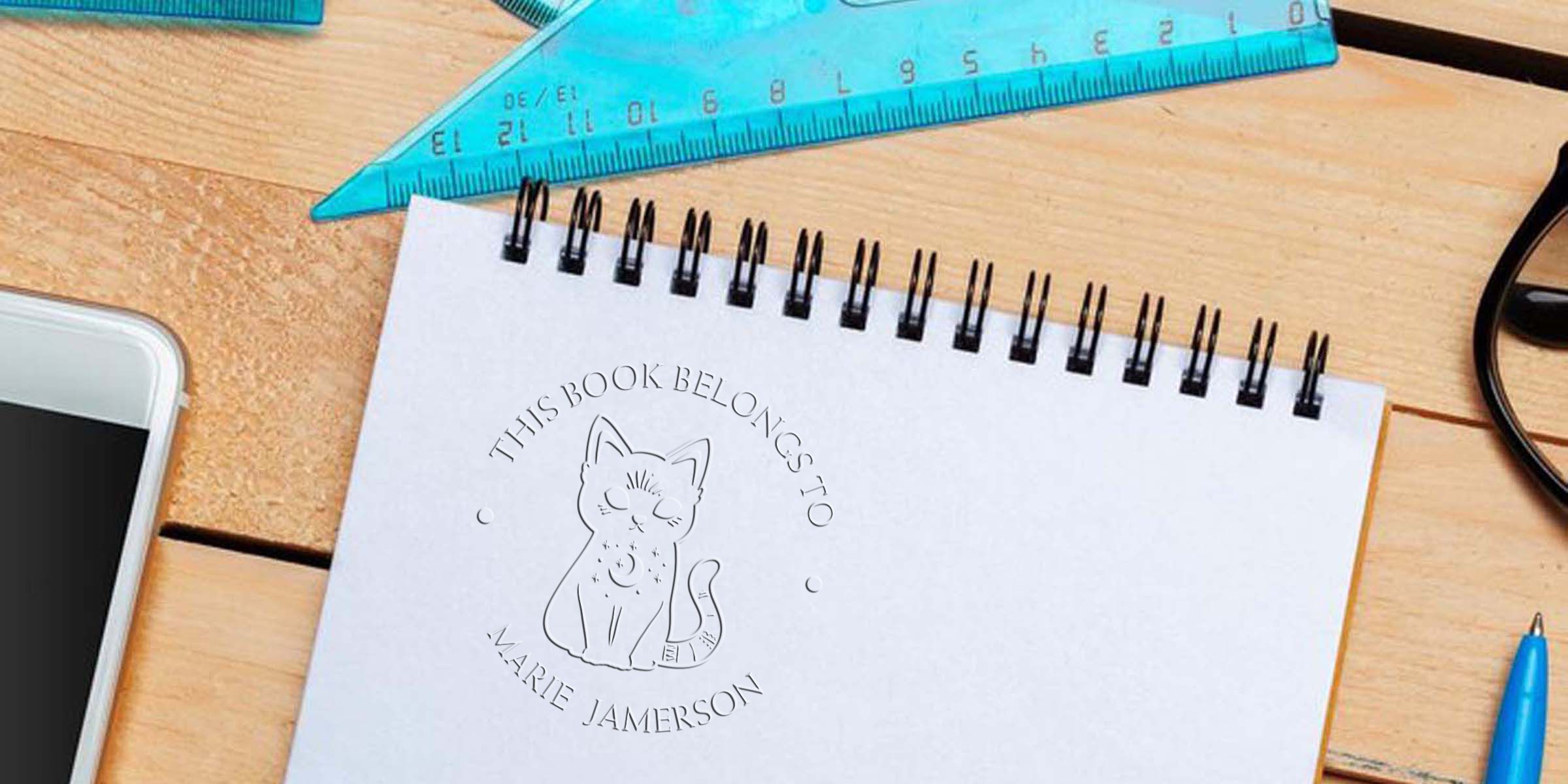 Spiral notebook with custom cat logo and text on a wooden desk, surrounded by a blue ruler, smartphone, and pen, ideal for stationery, branding, and school supplies themes