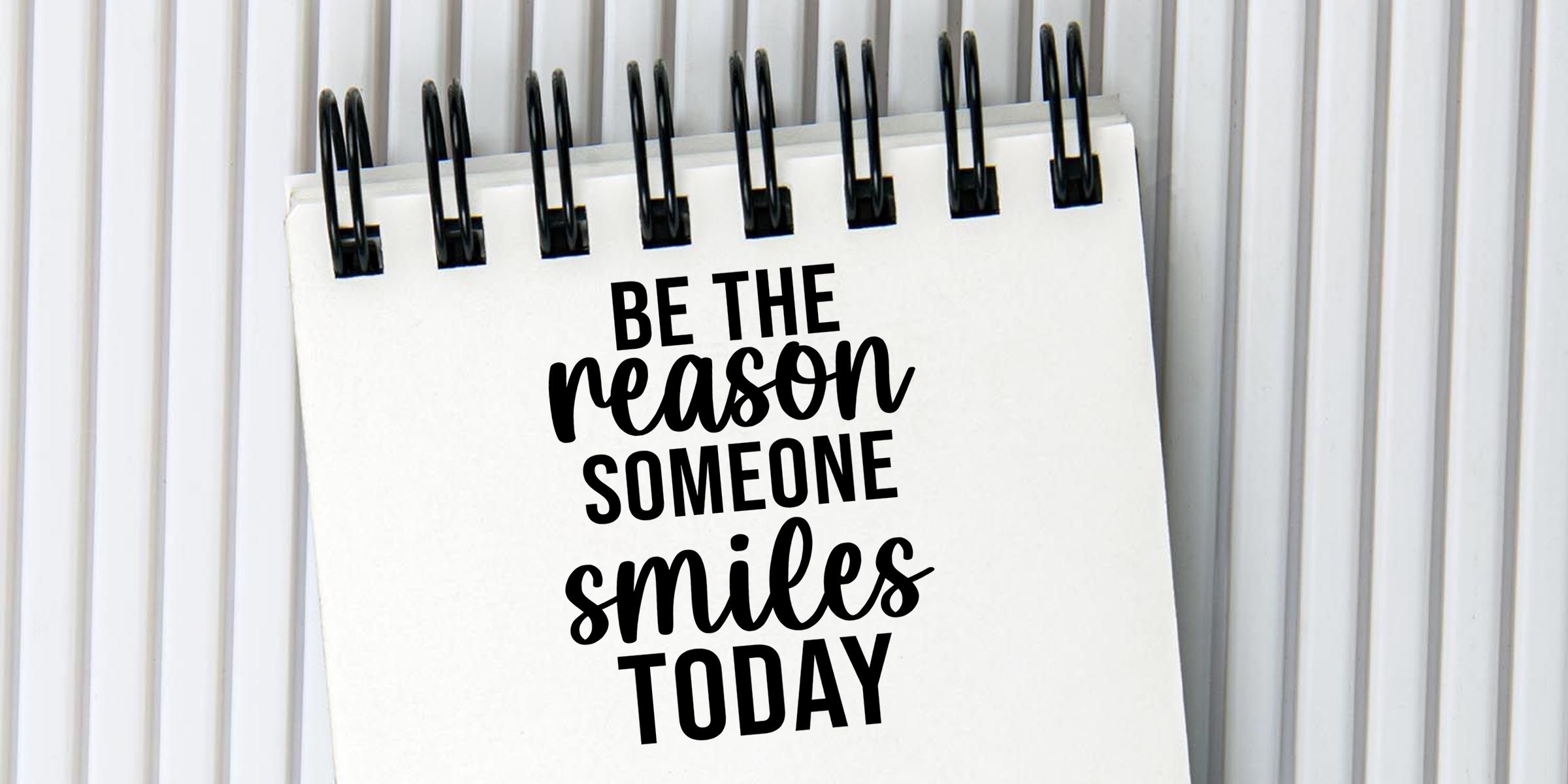 Spiral notebook with motivational quote “Be the reason someone smiles today” on a white page against a lined background