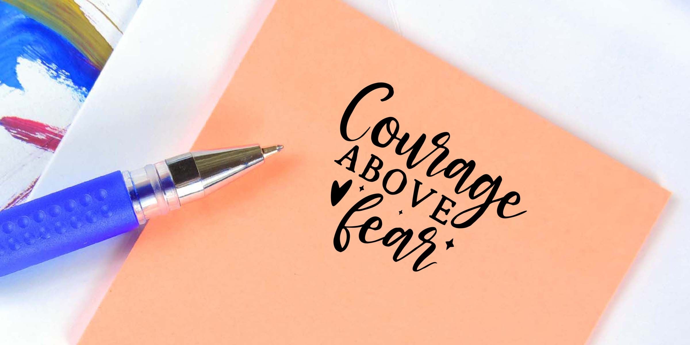 Blue ballpoint pen beside peach stationery with black calligraphy reading “Courage Above Fear” motivational quote design