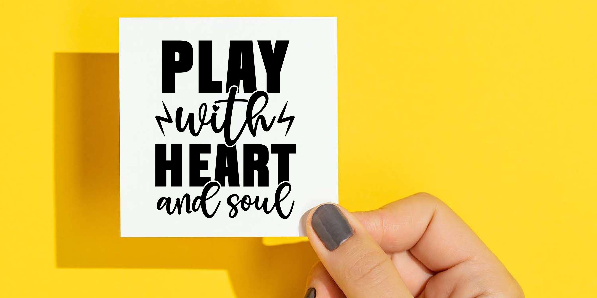 Hand holding a white card with the motivational quote “Play with heart and soul” in bold black typography on a bright yellow background