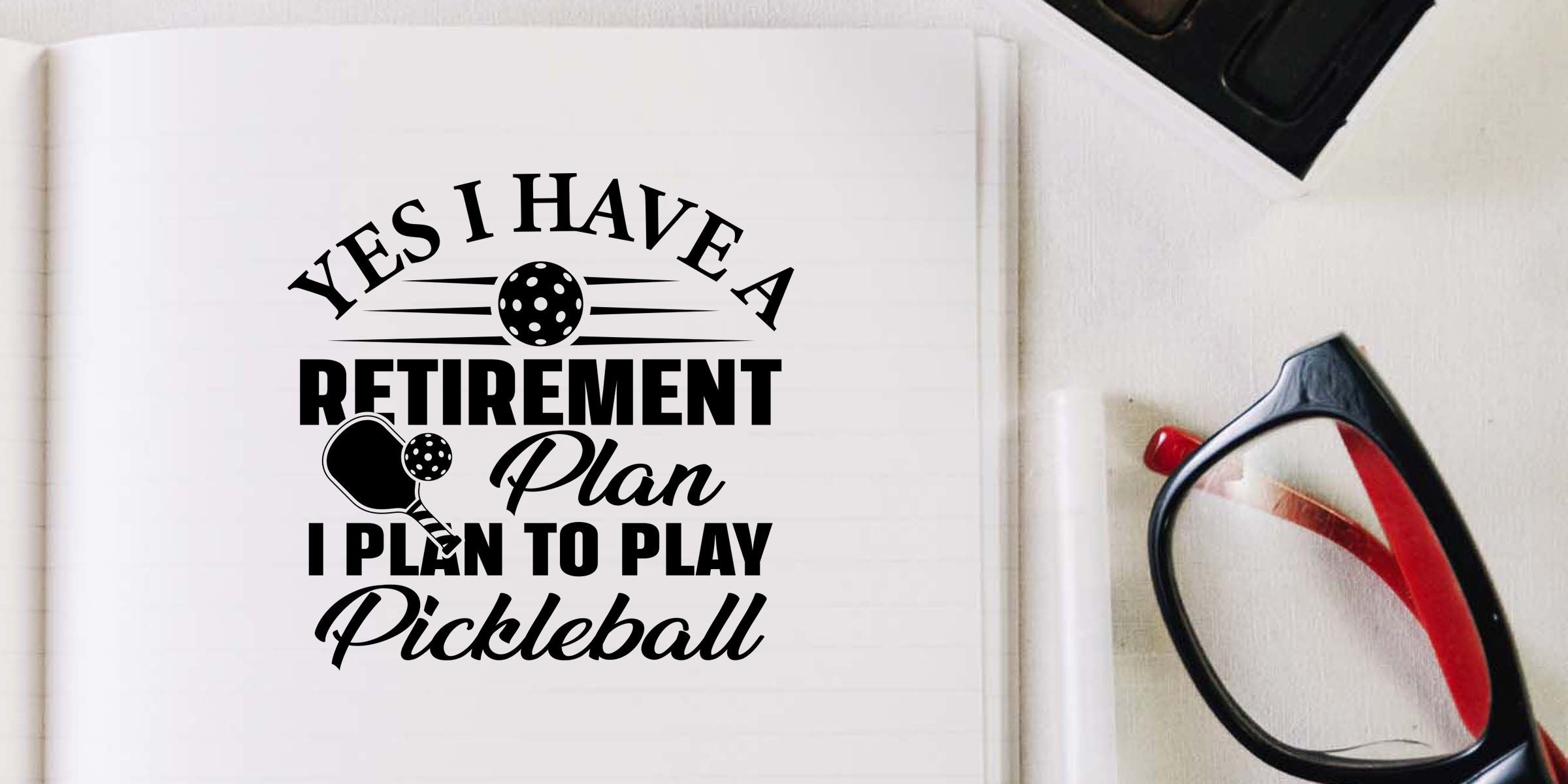 Notebook with “Yes I Have a Retirement Plan I Plan to Play Pickleball” quote, featuring a pickleball paddle, ball, and eyeglasses on a white desk