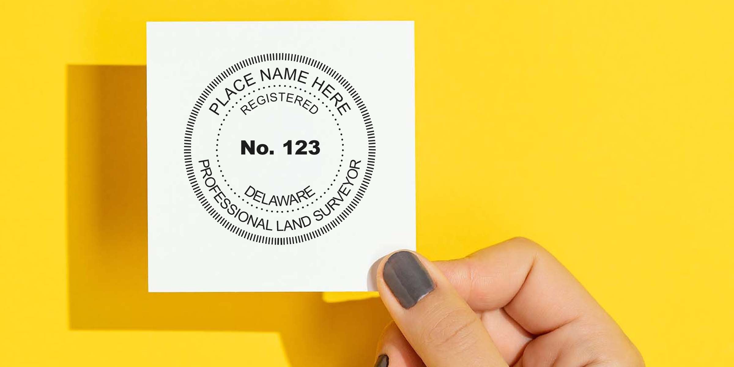Hand holding a Delaware land surveyor stamp with registration number on white paper against yellow background, highlighting Delaware land surveyor stamp cost and surveyor fees.