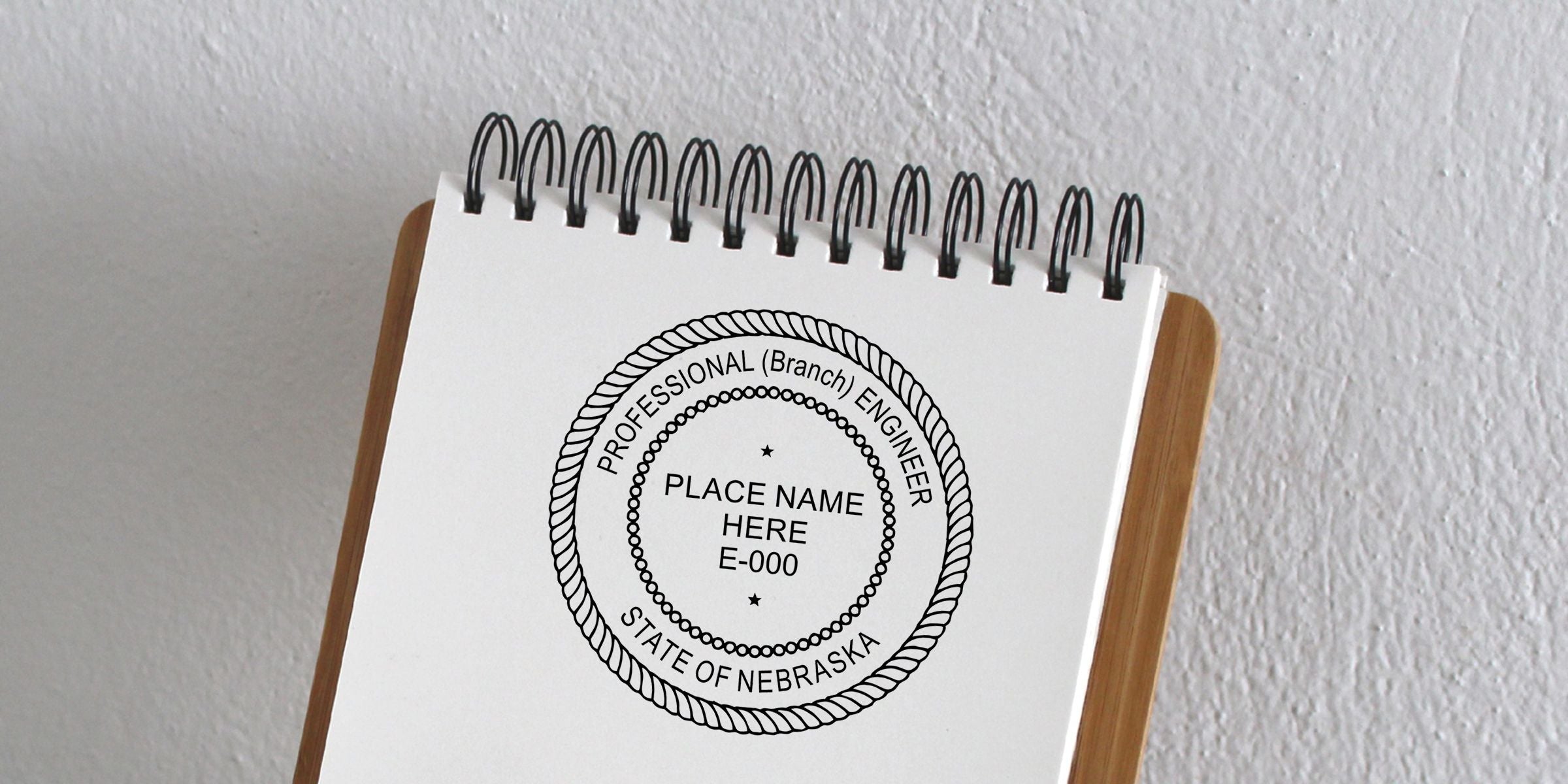 Spiral notebook displaying a Nebraska professional engineer seal template with place name here E-000 on a wooden stand against a white textured wall
