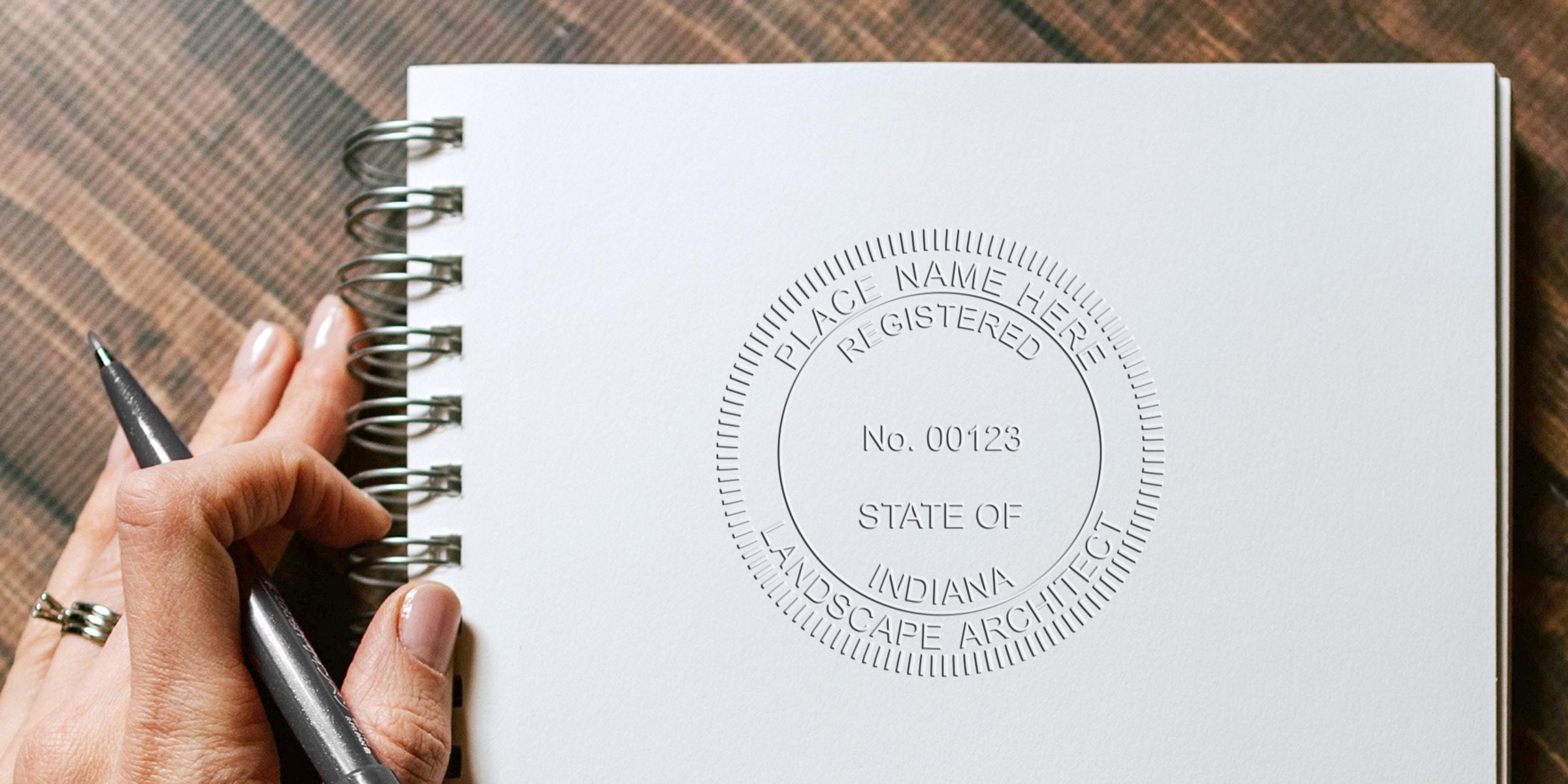 Hand holding pen next to embossed Indiana Landscape Architect Seal on notebook, highlighting the professional certification seal design.