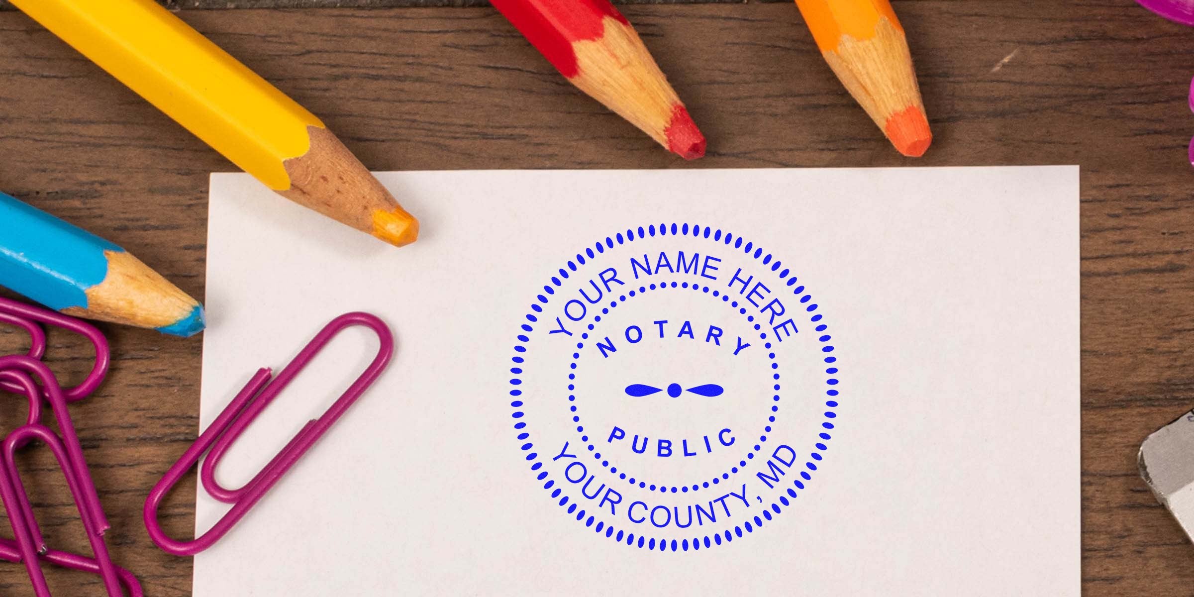 Maryland notary seal stamp on paper, surrounded by colorful pencils and paper clips on a wooden desk. Buy your notary seal online today.