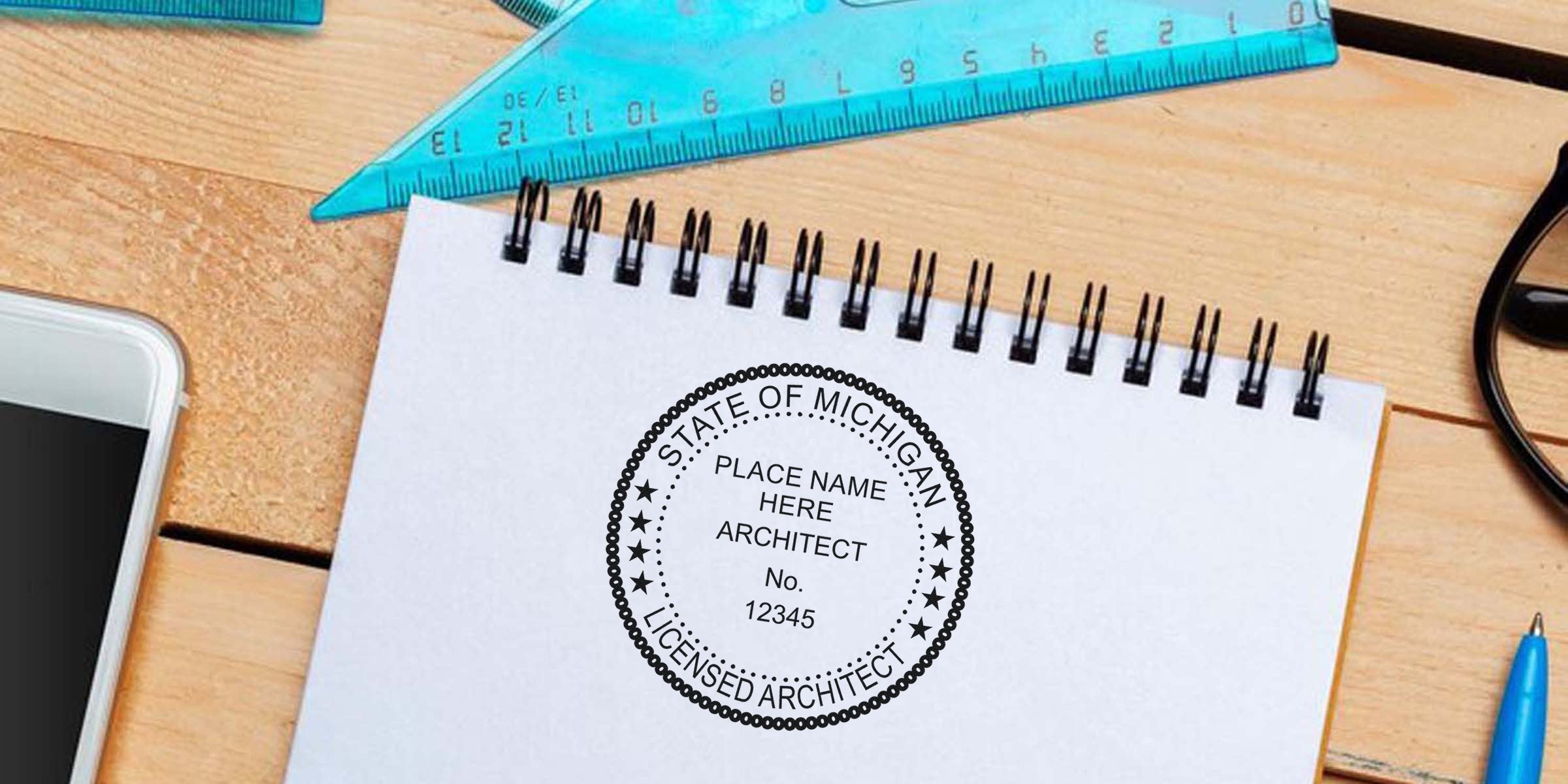 Michigan architect stamp on spiral notebook with ruler, smartphone, glasses, and pen on wooden desk, emphasizing Michigan architect stamp regulations.