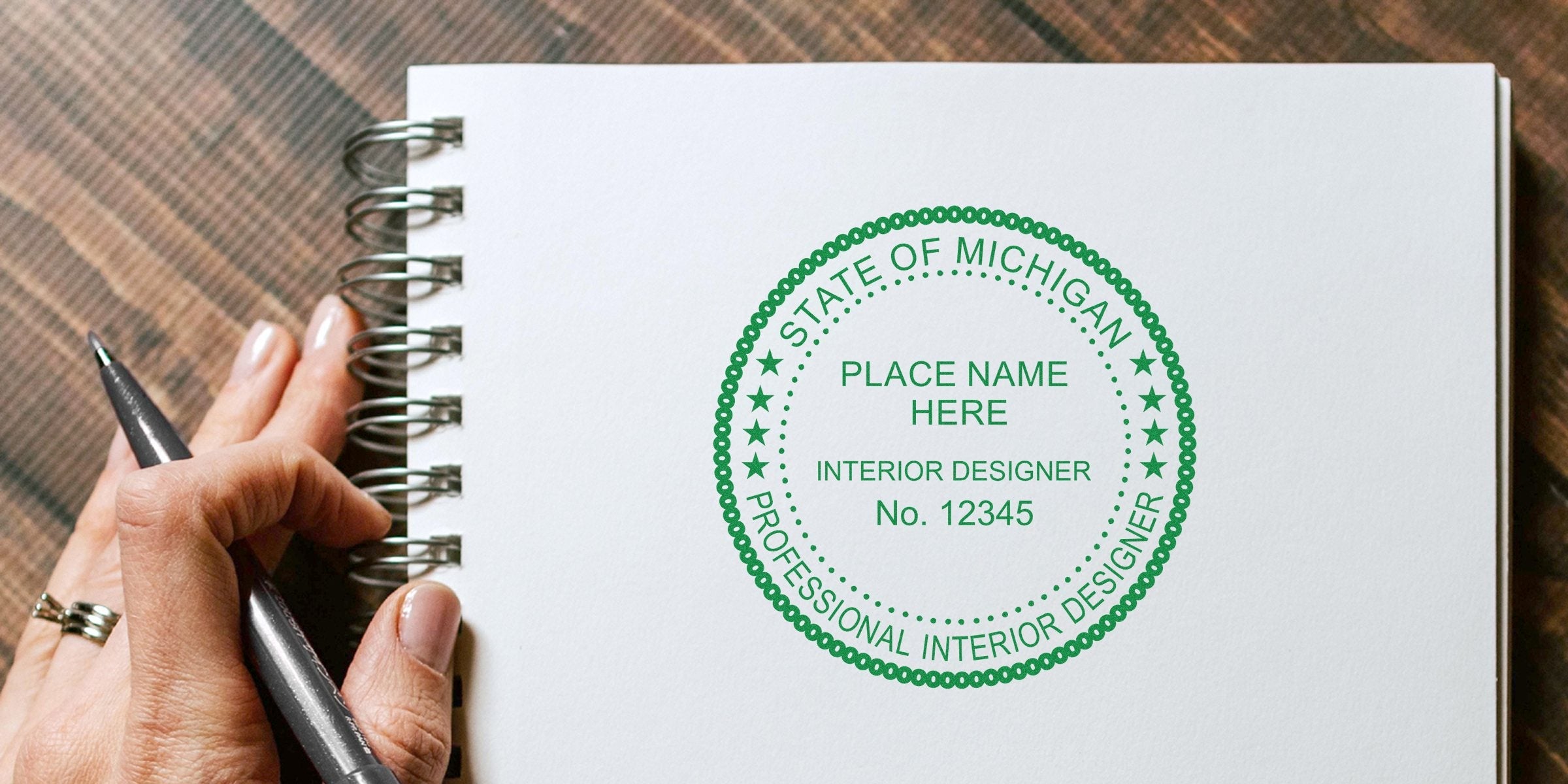 Hand holding pen next to Michigan interior designer seal stamp on notebook, showing stamp size for license verification.