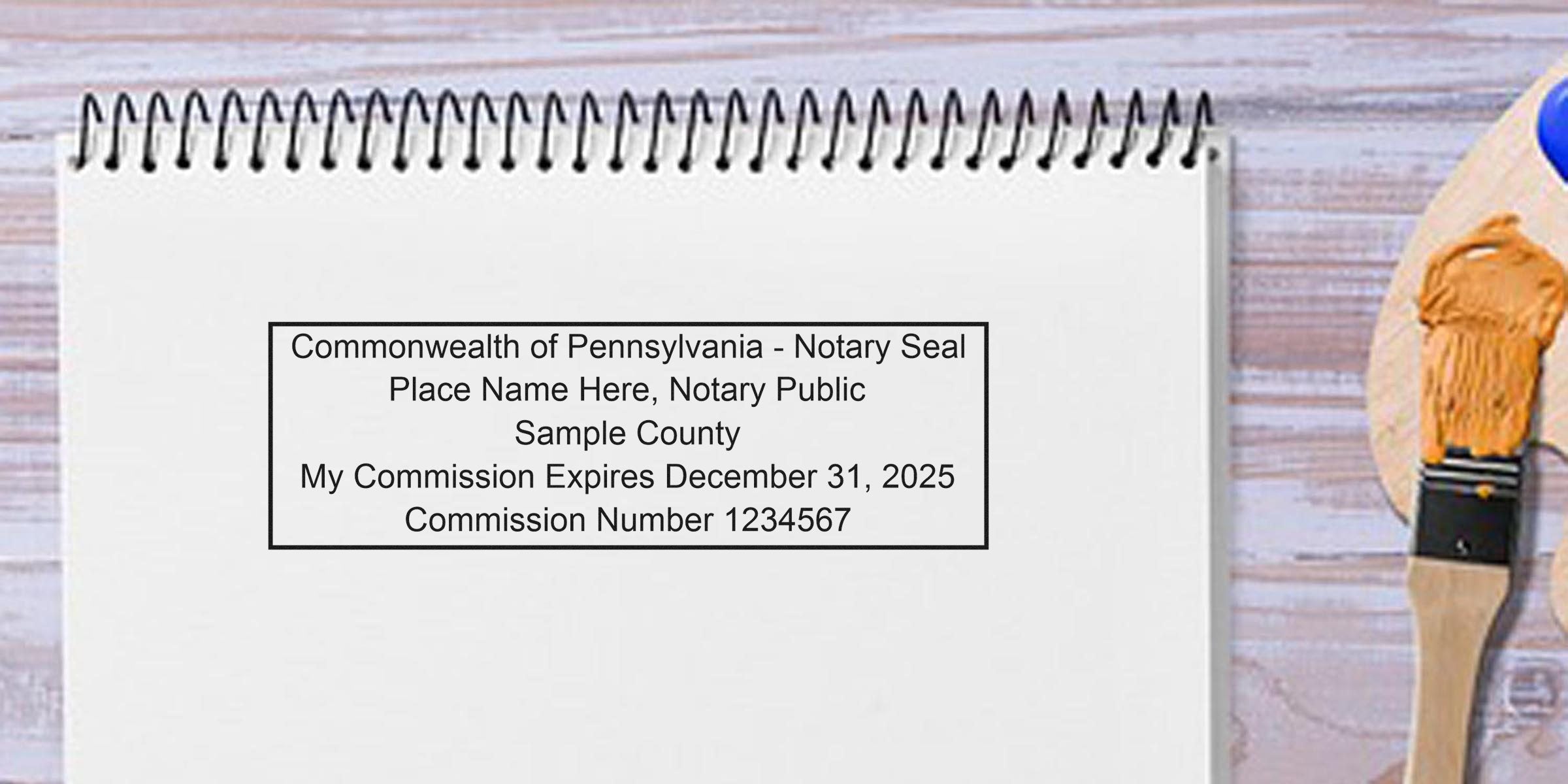 Commonwealth of Pennsylvania notary seal on white spiral notebook with paintbrush and palette on wooden table, showing seal size, dimensions, commission number, and expiration date.
