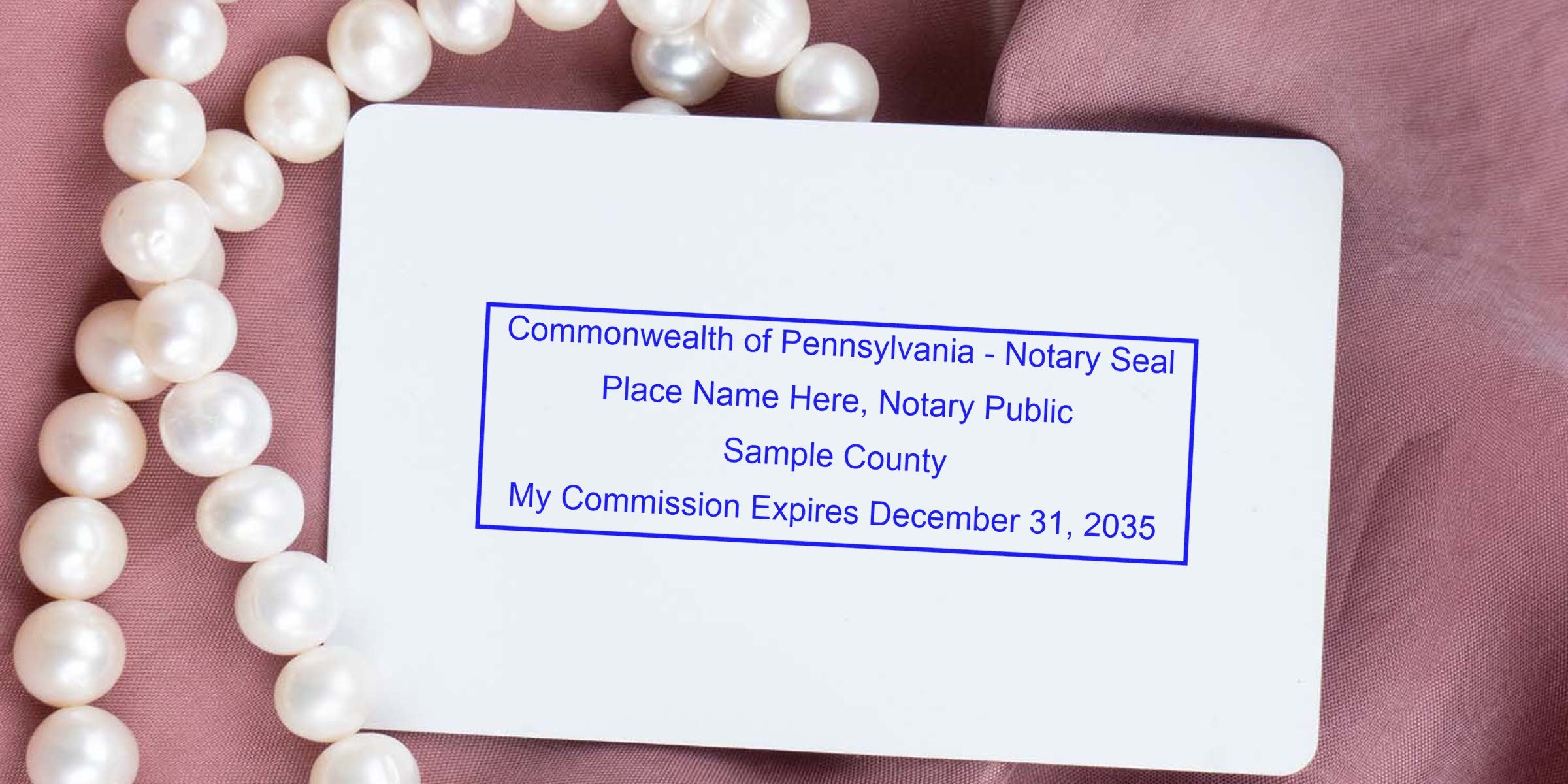 Pennsylvania notary seal sample on white card with commission expiration date, placed on pink fabric with pearl necklace, adhering to Pennsylvania notary seal guidelines.