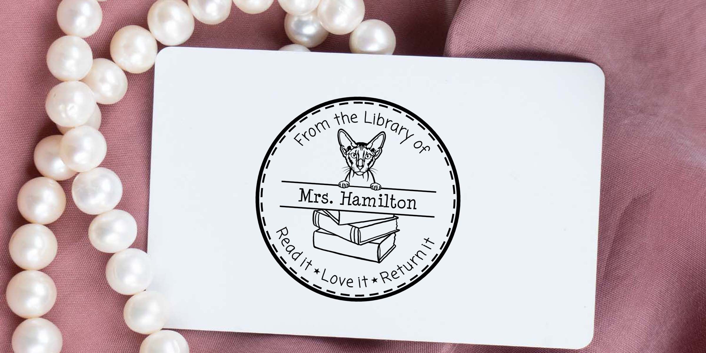 Personalized library bookplate sticker for Mrs. Hamilton with cat and books design, “Read it Love it Return it” text, displayed on pink fabric with pearl necklace.