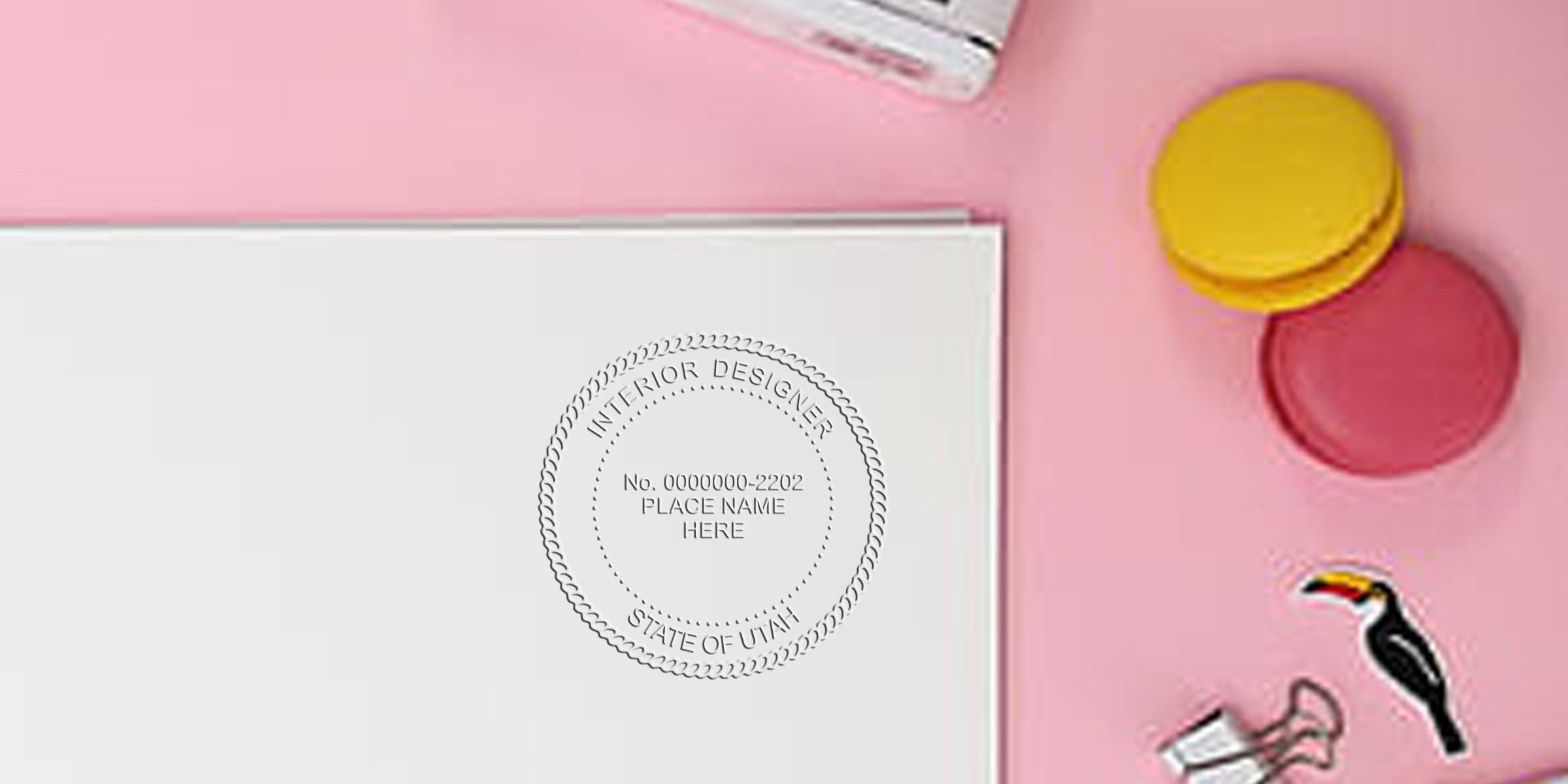 Utah interior designer stamp on white paper with pink and yellow macarons, binder clip, and toucan sticker on a pink background, meeting Utah interior design stamp requirements.