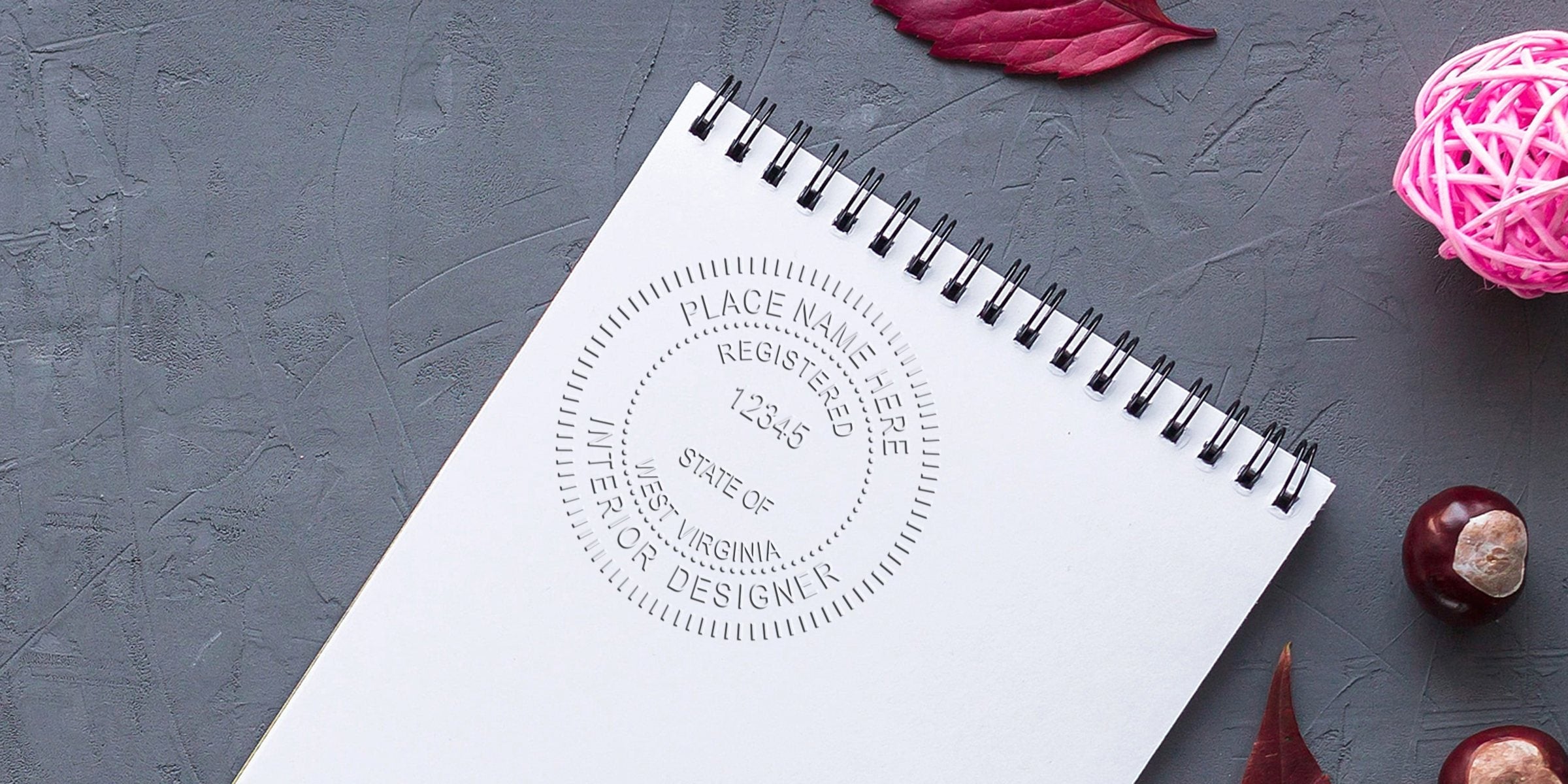 West Virginia interior designer stamp on white spiral notebook with autumn leaves and pink decorative ball on gray textured background, highlighting the WV interior design licensing process.