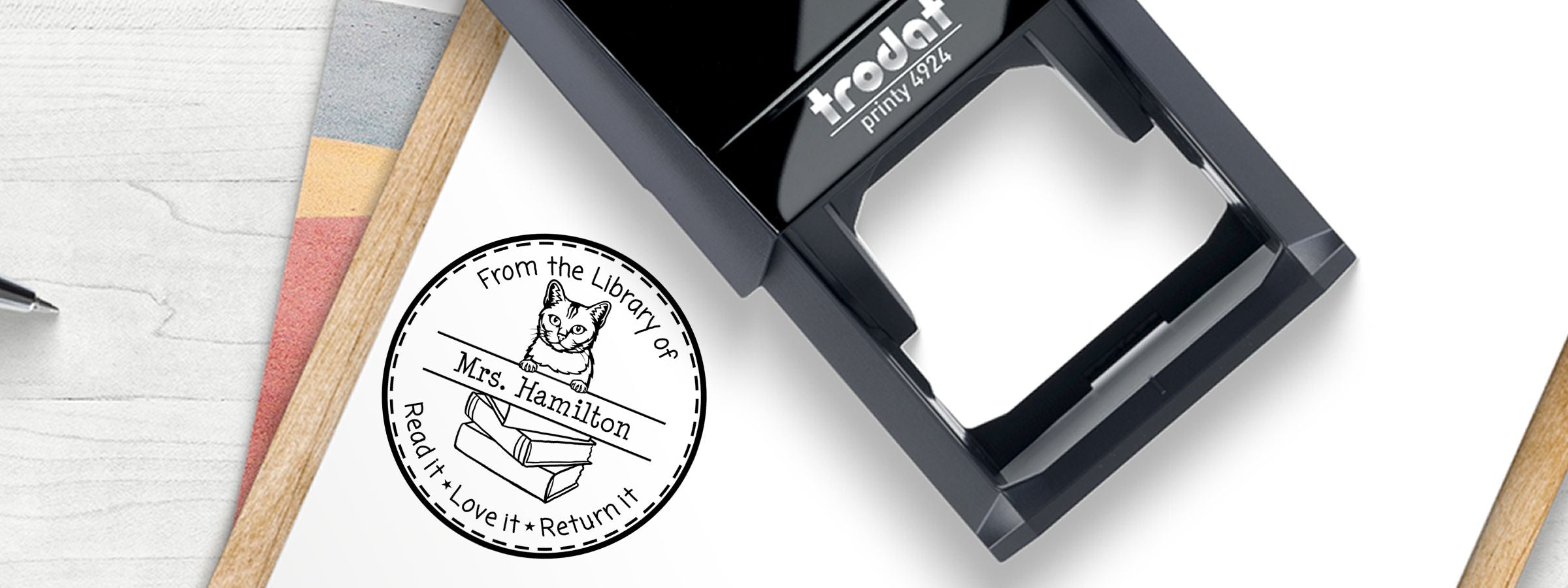 Burmilla Cat Stamps and Custom Seals self-inking library return stamp with cat and books, beside a black Trodat case.