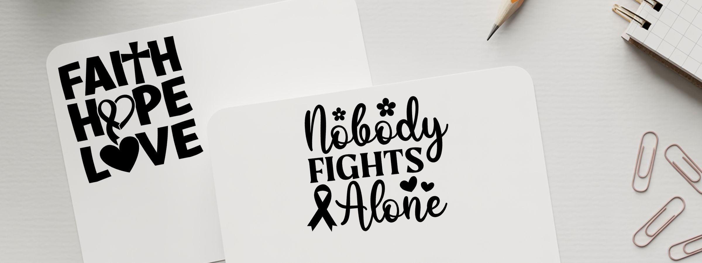 Cancer Awareness Craft Stamps on two white cards reading Faith Hope Love and Nobody Fights Alone, with hearts and ribbon on a desk.