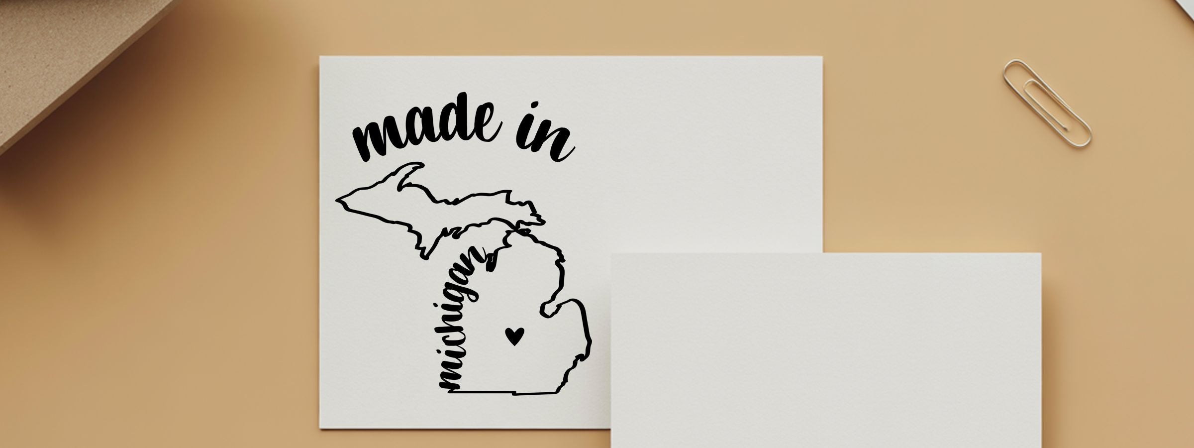 White stationery mockup on tan background, card reads “made in Michigan” with state outline heart and paper clip; Michigan Designer Stamps and Seals.