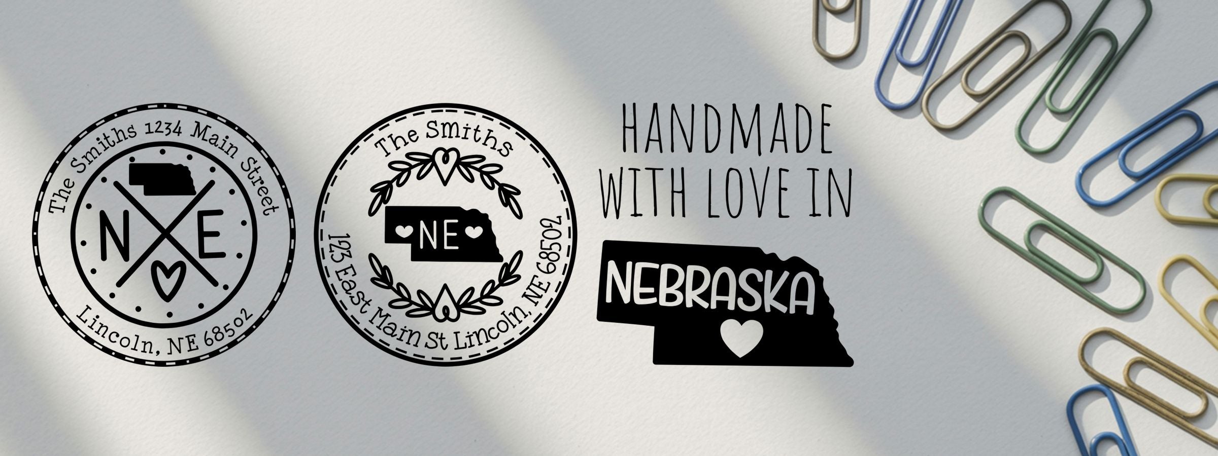 Three black Nebraska-themed handmade design logos on white paper, with colorful paper clips along the right edge, for Nebraska Designer Stamps and Seals.