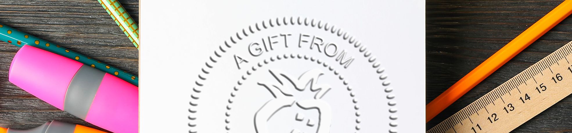 White paper embossed with “A GIFT FROM” and fruit logo, with colorful pens, pencil, and ruler on a dark desk; Library / Book Embossers.