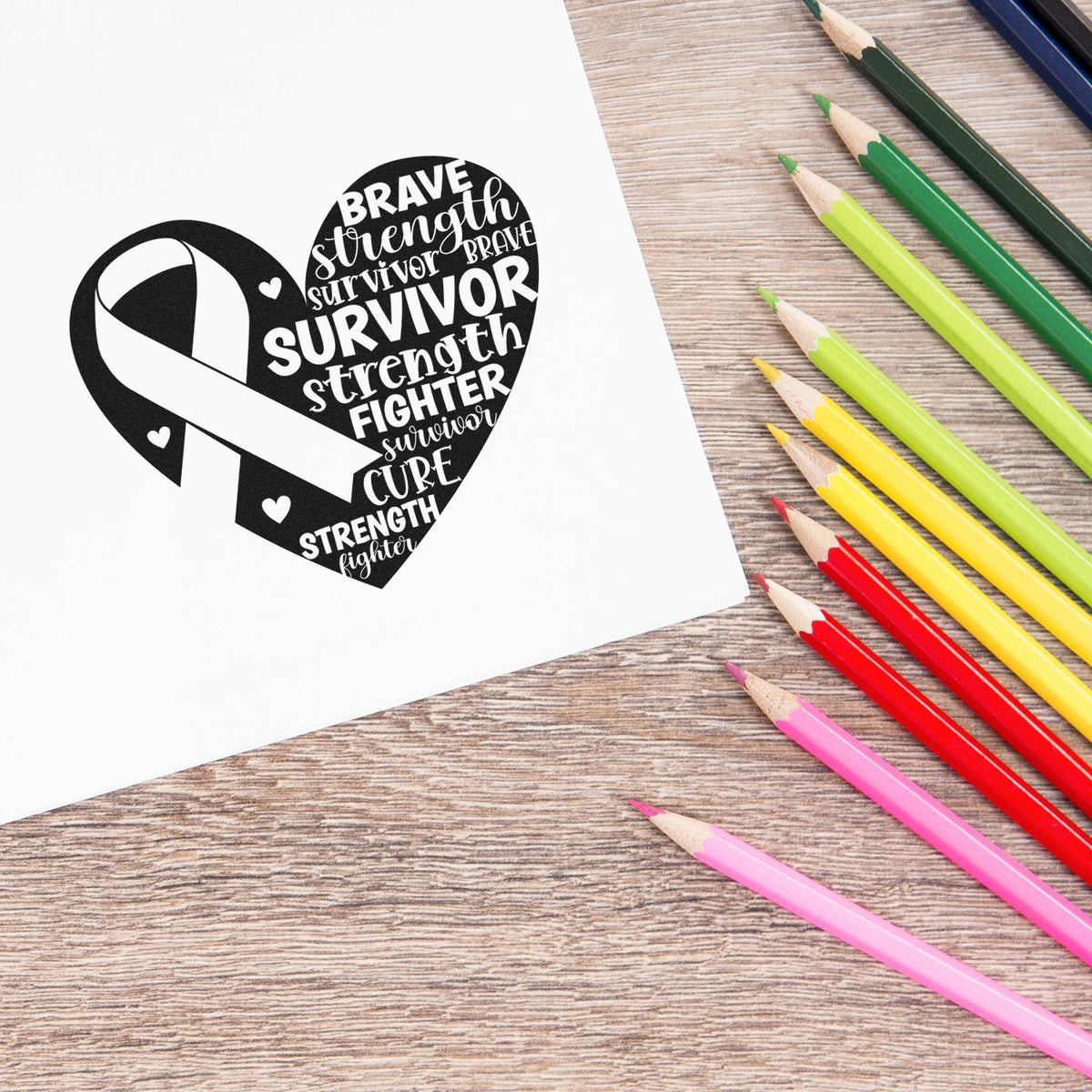 Breast cancer awareness rubber stamp design with heart ribbon and survivor words, shown beside colored pencils on white paper.