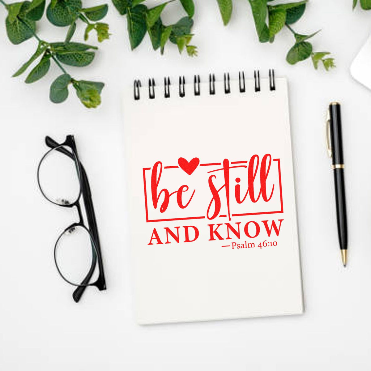Christian rubber stamp design with Be Still and Know Psalm 46:10 on a notebook, styled on a desk with pen and glasses.