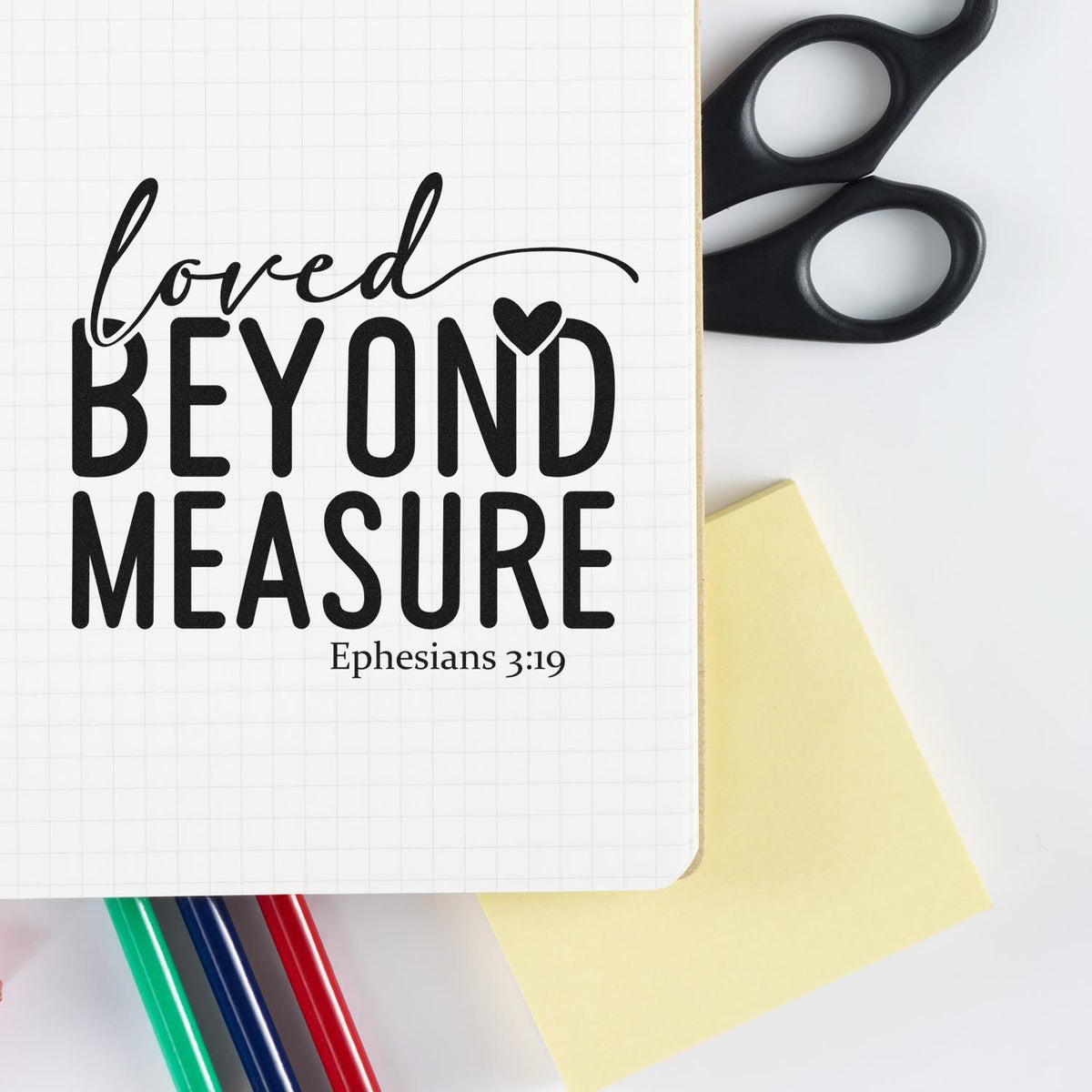 Christian rubber stamp design reading Loved Beyond Measure with Ephesians 3:19, shown on a notebook with craft supplies.