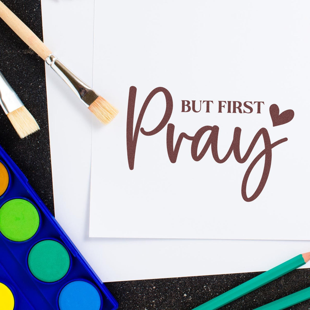Rubber stamp design reading But First Pray on paper with paintbrushes and watercolor palette for DIY crafting.