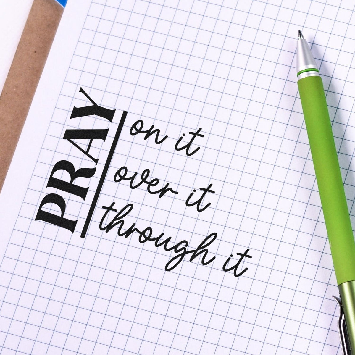 Christian prayer rubber stamp design with Pray on it, over it, through it on graph paper beside a green pen.