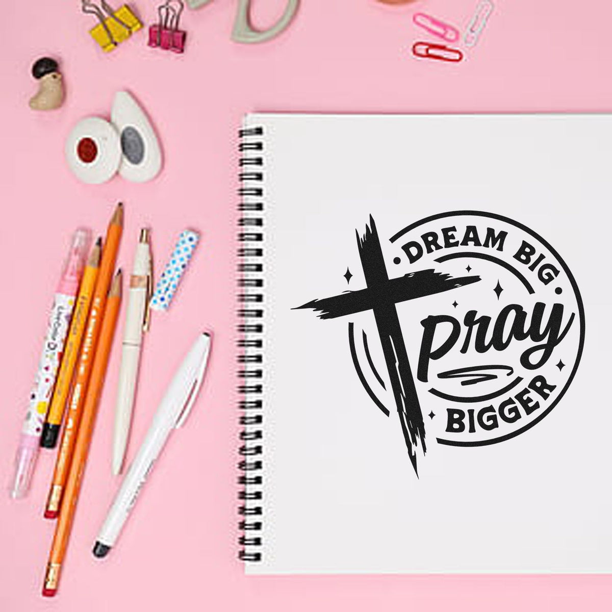 Dream Big Pray Bigger rubber stamp design on a spiral notebook with pens on a pink desk, perfect for inspirational crafting.