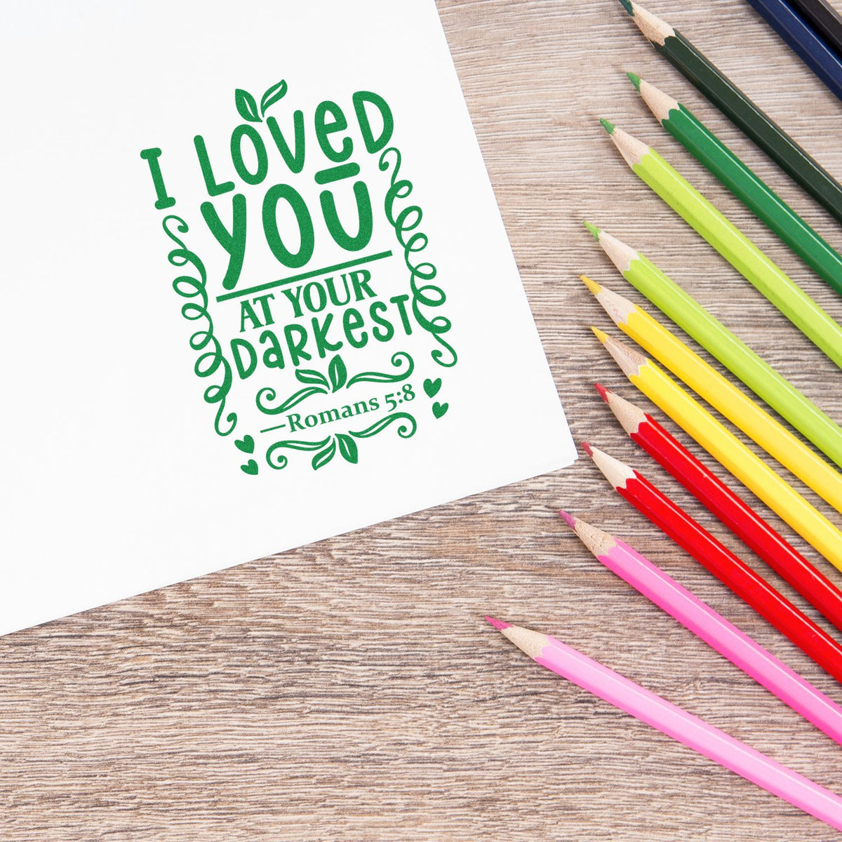 Romans 5:8 Christian rubber stamp design reading I loved you at your darkest with colorful pencils on a desk.