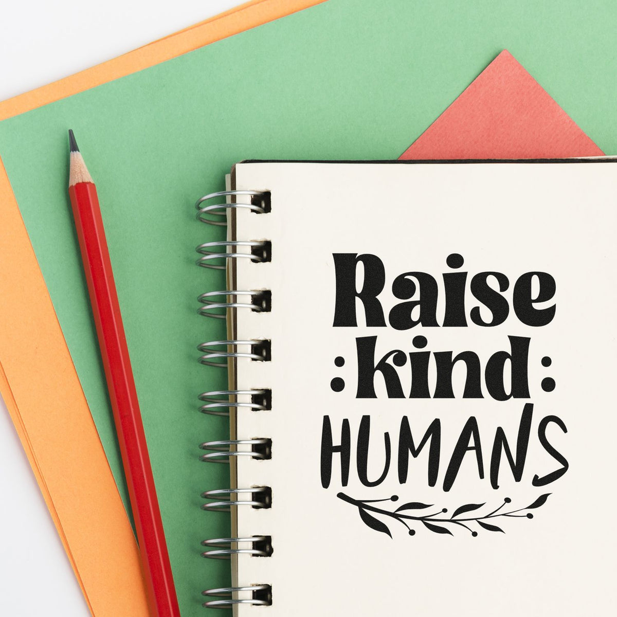 Raise Kind Humans rubber stamp design on a notebook page with pencil and colorful paper background.