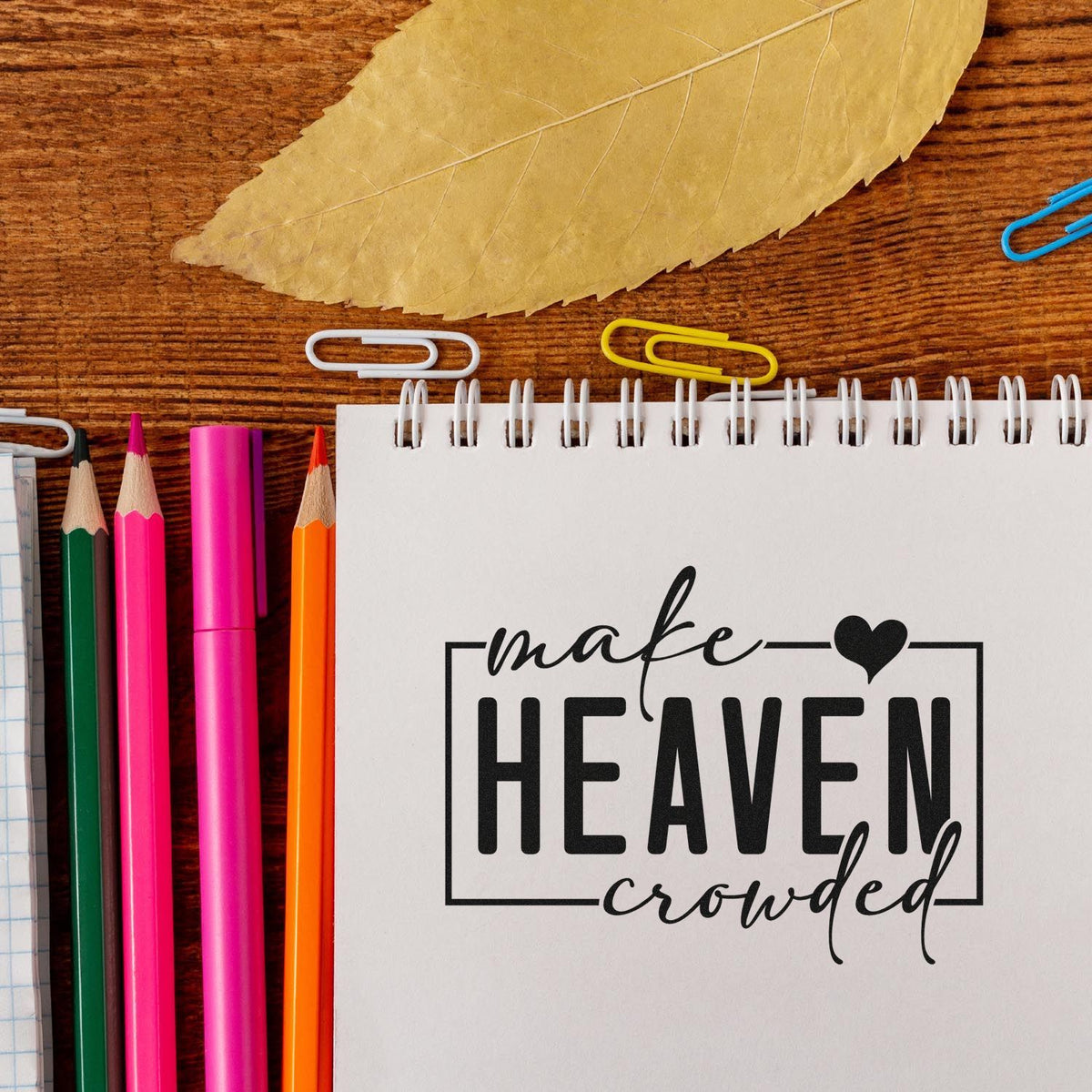 Inspirational rubber stamp design reading make heaven crowded on a notebook with colorful pencils and paper clips.