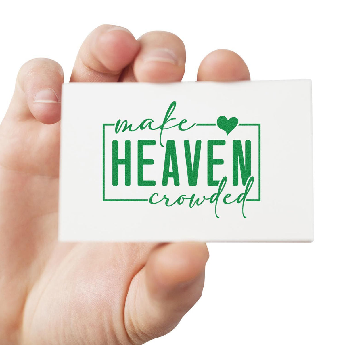 Hand holding Christian rubber stamp reading make heaven crowded in green script and bold lettering with heart design.