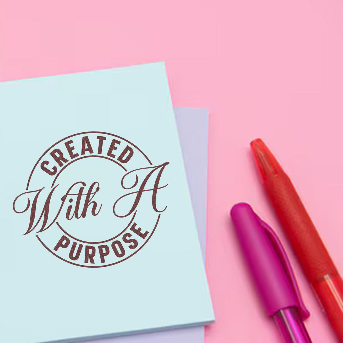 Custom rubber stamp reading "Created With A Purpose" on pastel paper with pens for branding and packaging.