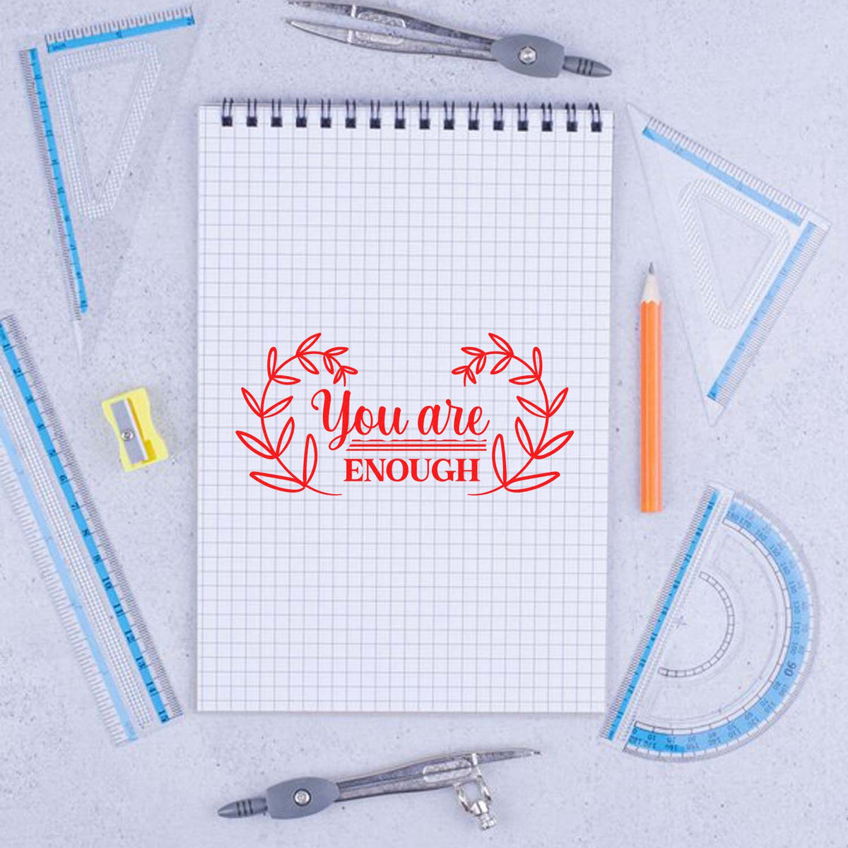 Inspirational rubber stamp design with You are enough and leafy wreath, shown on sketchpad with drafting tools.
