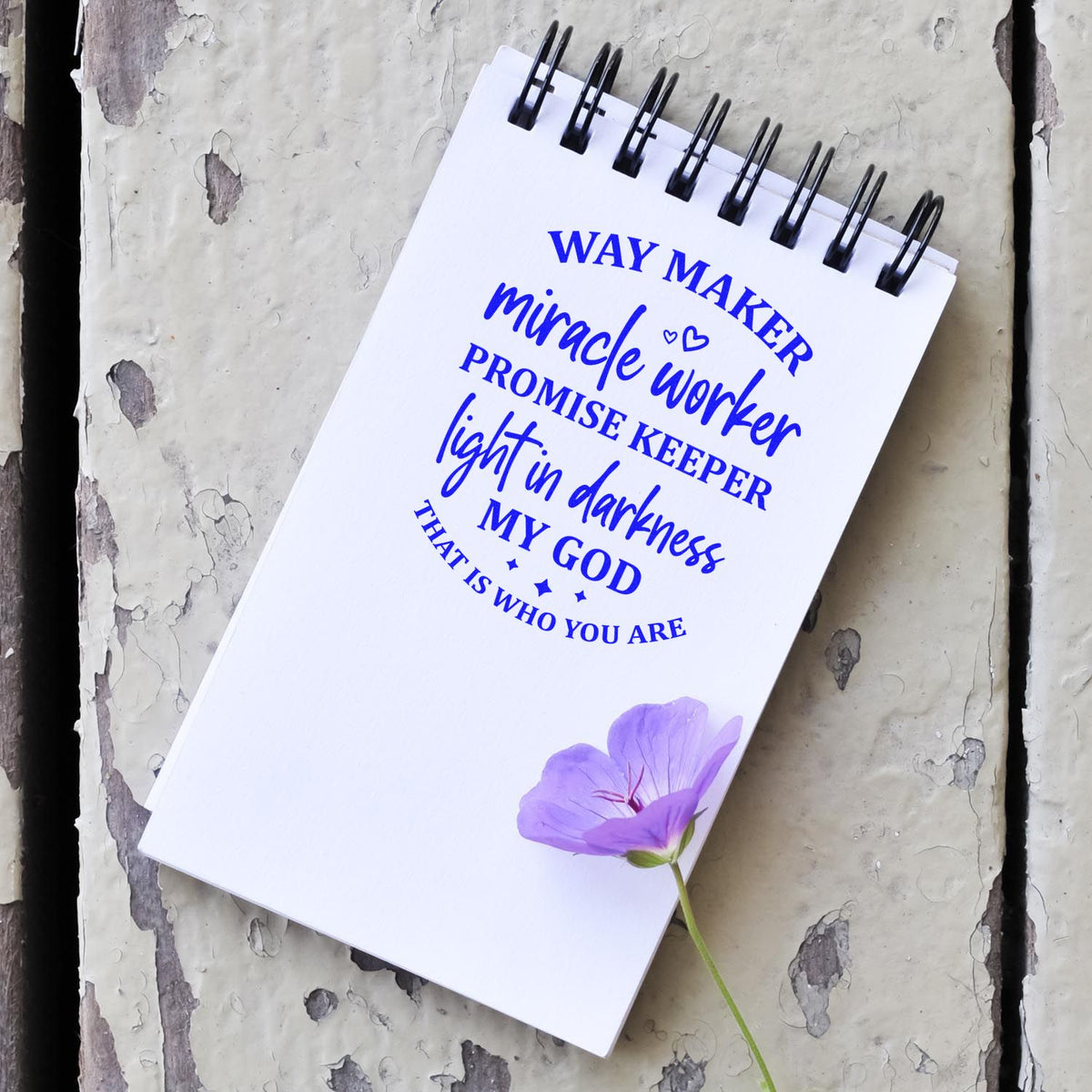 Blue inspirational rubber stamp on notebook reading Way Maker, Miracle Worker, Promise Keeper, with purple flower accent.