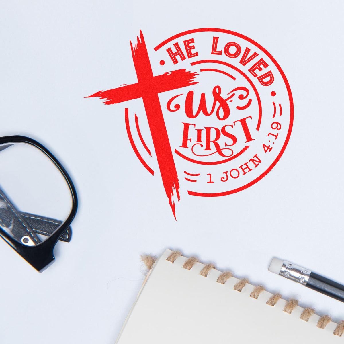 Red Christian rubber stamp imprint with cross and Bible verse He Loved Us First from 1 John 4:19.