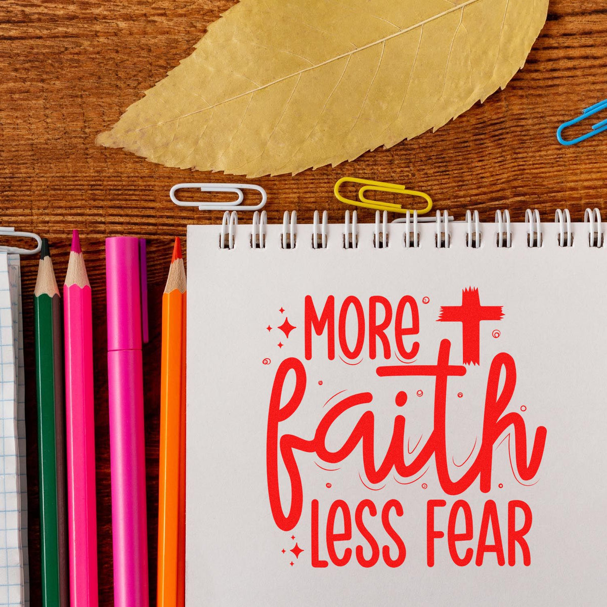 Inspirational rubber stamp design with More Faith Less Fear on notebook, surrounded by colorful pencils and paper clips.