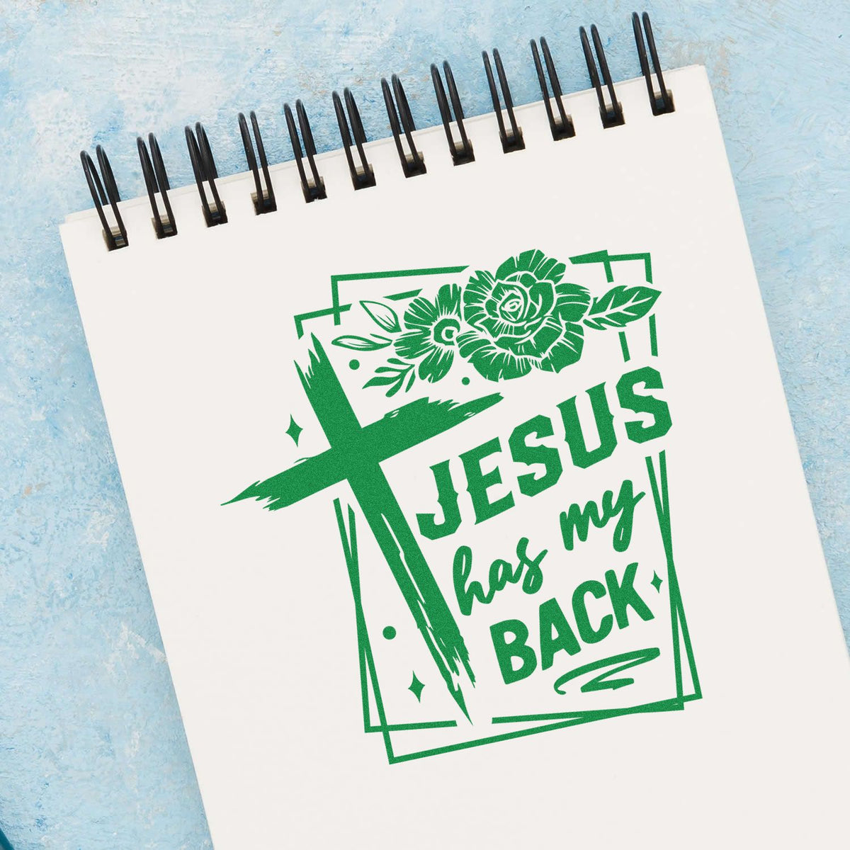 Faith-based rubber stamp design reading Jesus has my back with cross and floral accents on a notebook page.