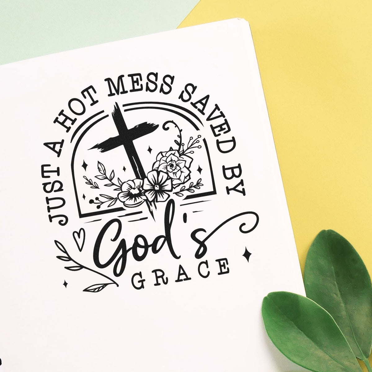 Just A Hot Mess Saved By God's Grace Slim Pre-Inked Stamp imprint stamped in black ink. A floral cross sits inside an arch surrounded by "Just a hot mess saved by God's grace."