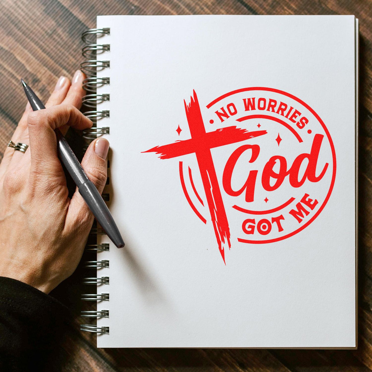 Hand beside notebook with red Christian rubber stamp design reading No Worries God Got Me with cross graphic.