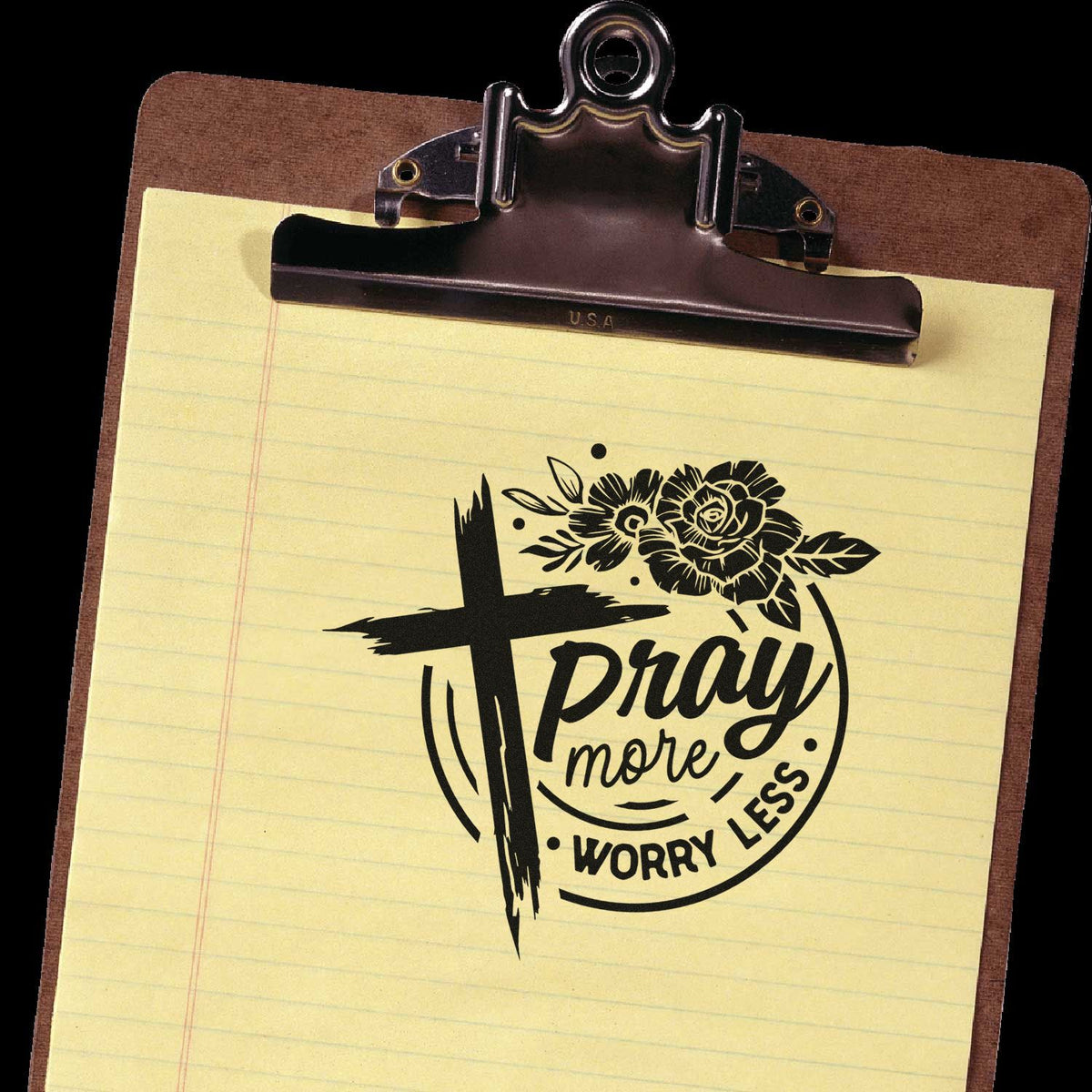 Christian rubber stamp design with cross and flowers reading Pray more, worry less on lined paper clipboard.