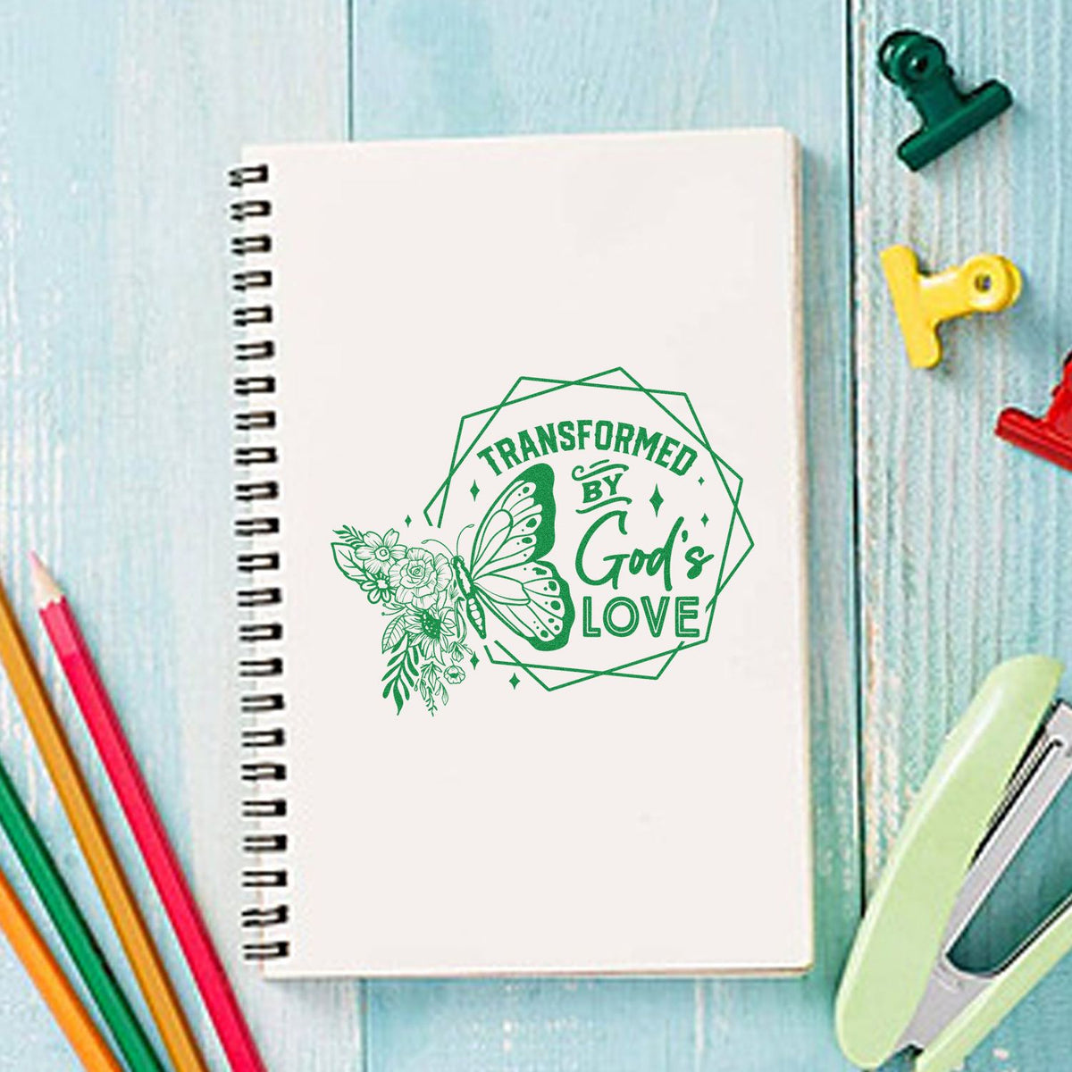 Faith-based butterfly floral rubber stamp design reading Transformed by Gods Love on a spiral notebook.