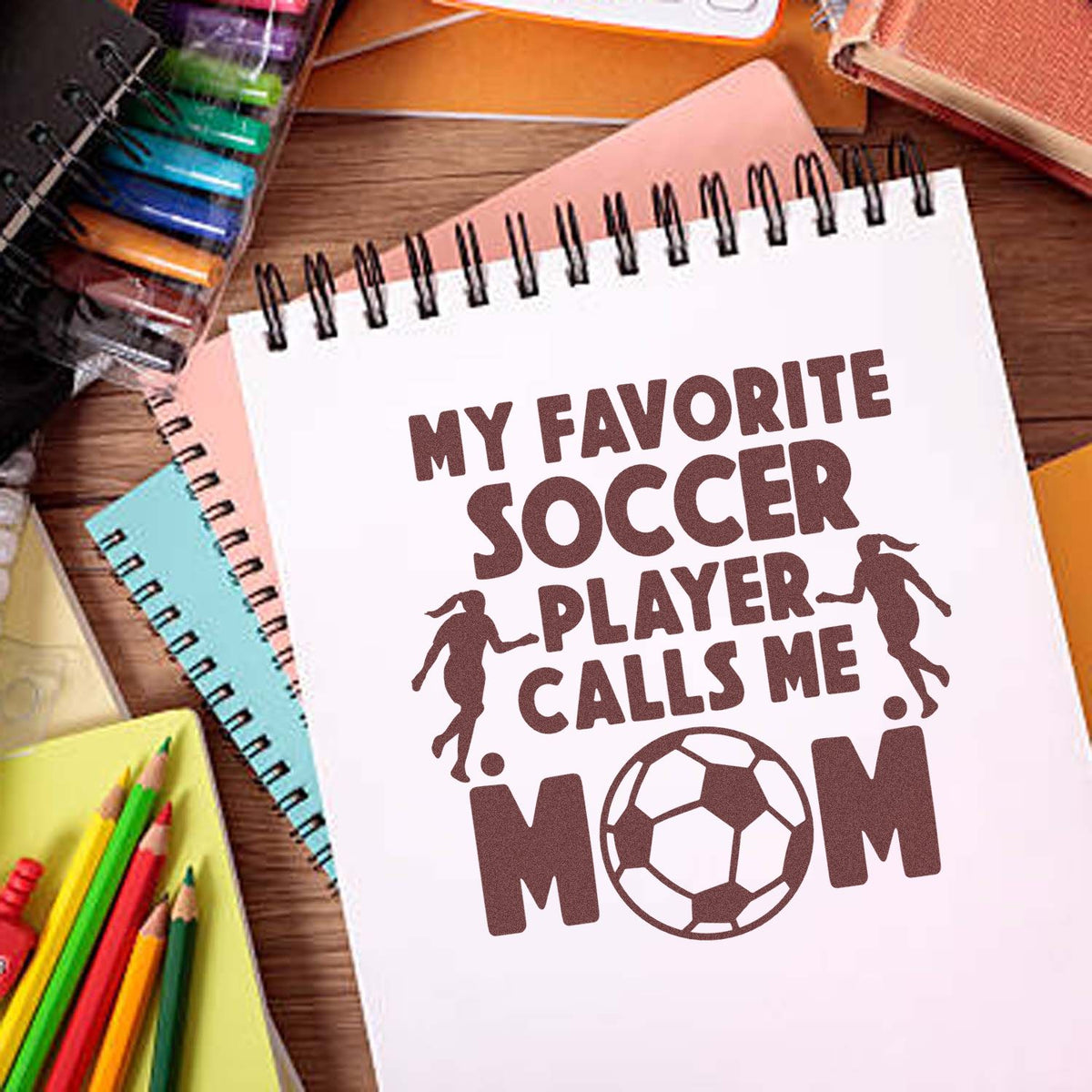 Soccer mom rubber stamp design with quote "My favorite soccer player calls me mom" and soccer ball graphic on notebook.